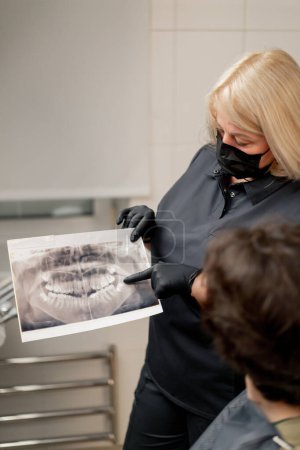 Foto de Odontología rubia odontóloga en uniforme negro consultando a un paciente mostrando una foto de los dientes - Imagen libre de derechos