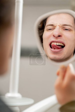 Téléchargez les photos : Fermer le cabinet dentaire jeune homme assis et regarde ses dents à travers un miroir dentaire en utilisant sa langue sur ses dents - en image libre de droit