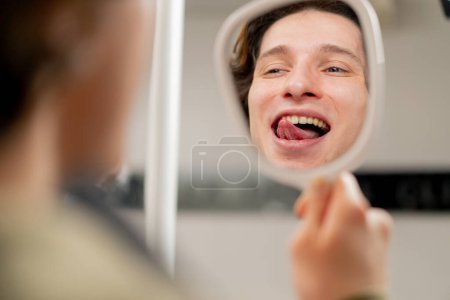 Téléchargez les photos : Fermer le cabinet dentaire jeune homme assis et regarde ses dents à travers un miroir dentaire en utilisant sa langue sur ses dents - en image libre de droit