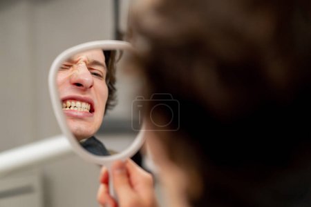 Téléchargez les photos : Fermer le cabinet dentaire un jeune homme assis dans une chaise et regarde ses dents à travers un miroir dentaire - en image libre de droit