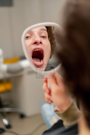 Photo for Close up dental office a young guy sits in a chair and looks at his teeth through a dental mirror open mouth - Royalty Free Image