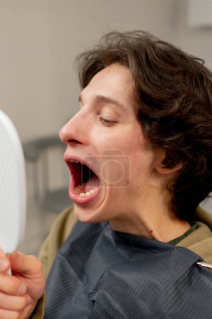 Téléchargez les photos : Fermer le cabinet dentaire un jeune homme assis dans une chaise et regarde ses dents à travers un miroir dentaire bouche ouverte - en image libre de droit