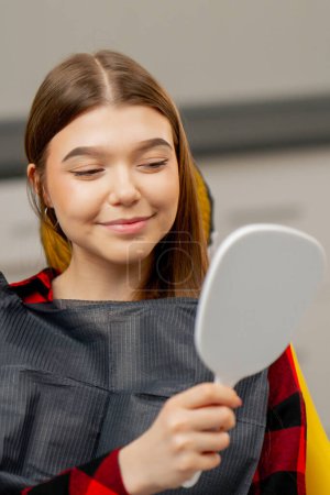 Photo for Dental office a young girl sits and looks at her teeth in a dental mirror rejoicing - Royalty Free Image