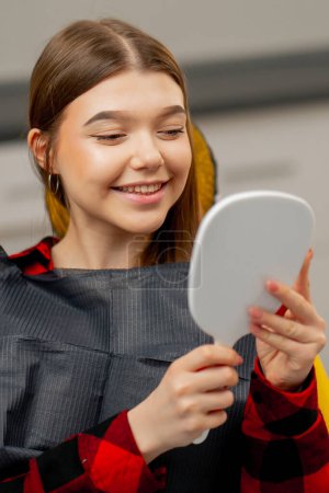 Téléchargez les photos : Cabinet dentaire une jeune fille s'assoit et regarde ses dents dans un miroir dentaire se réjouissant - en image libre de droit