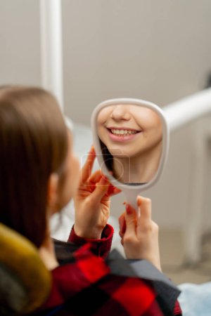 Téléchargez les photos : Cabinet dentaire une jeune fille s'assoit et regarde ses dents à travers un miroir dentaire - en image libre de droit