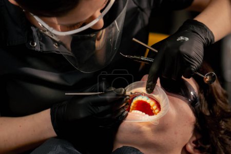 Téléchargez les photos : Dentiste fille de bureau dentaire applique gel protecteur sur les gencives d'un jeune homme - en image libre de droit