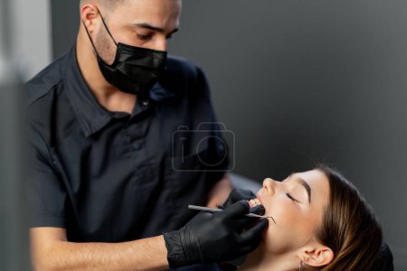 Foto de En un consultorio dental un dentista masculino examina los dientes de una joven hermosa - Imagen libre de derechos