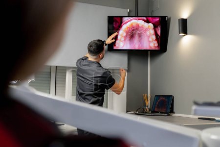 Foto de En un consultorio dental un hombre muestra una foto de los dientes en un monitor para una niña hermosa - Imagen libre de derechos