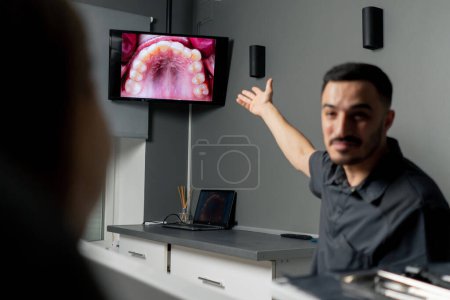 Foto de En un consultorio dental un hombre muestra una foto de los dientes en un monitor para una joven hermosa consulta de la muchacha - Imagen libre de derechos