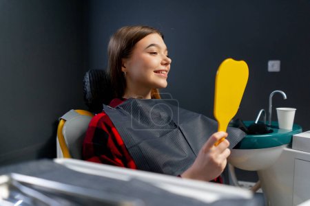 Photo for In the dental office young girl sits and looks at her teeth in a yellow dental mirror rejoicing - Royalty Free Image