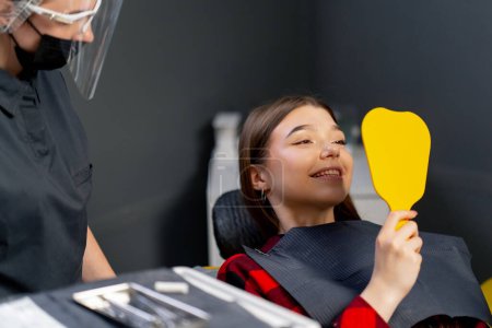 Photo for In the dental office young girl sits and looks at her teeth in a yellow dental mirror rejoicing - Royalty Free Image
