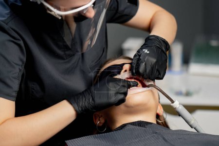 Foto de En un consultorio dental odontóloga con gorra hace hilo dental a una joven hermosa chica - Imagen libre de derechos