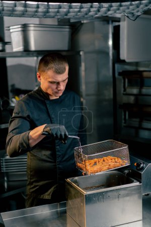 Photo for The chef in the kitchen in a black jacket takes out a baht from the deep fryer - Royalty Free Image