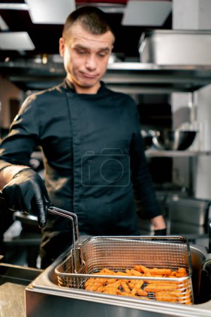 Photo for The chef in the kitchen in black jacket stands and holds a fryer and thoughtfully looks at the sweet potato close-up - Royalty Free Image