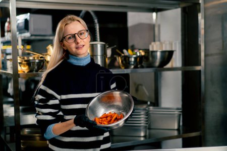 Foto de Una joven con gafas en la cocina se para con un tazón de batata demostrando mirar a la cámara - Imagen libre de derechos