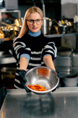 Photo for A young girl with glasses in the kitchen stands and holds out a bowl of sweet potato demonstrating the product to the camera - Royalty Free Image