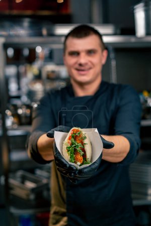 Téléchargez les photos : Gros plan d'un chef cuisinier dans la cuisine d'un restaurant vêtu d'une veste noire démontrant un petit pain prêt à l'emploi aux crevettes - en image libre de droit