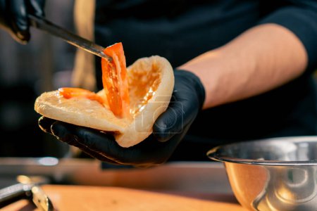 Foto de Primer plano chef en guantes negros pone diferentes ingredientes en un bollo con pinzas - Imagen libre de derechos
