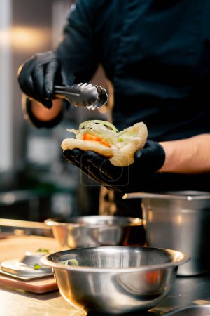 Foto de Primer plano chef en guantes negros pone diferentes ingredientes en un bollo con pinzas - Imagen libre de derechos