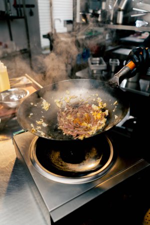 Foto de Primer plano de una sartén con huevo y carne sobre la estufa con una mano en un tiro para mezclar el contenido - Imagen libre de derechos