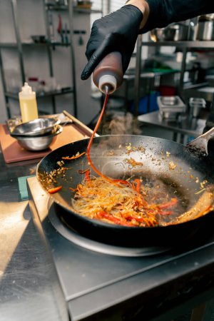 Foto de Primer plano de sartén con fideos huevos y carne en la estufa el chef vierte la salsa - Imagen libre de derechos