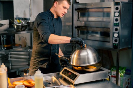 a chef in a restaurants kitchen in a black jacket opens the lid of a frying pan to check its readiness