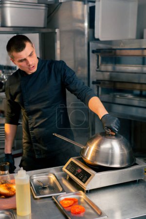 a chef in a restaurants kitchen in a black jacket opens the lid of a frying pan to check its readiness