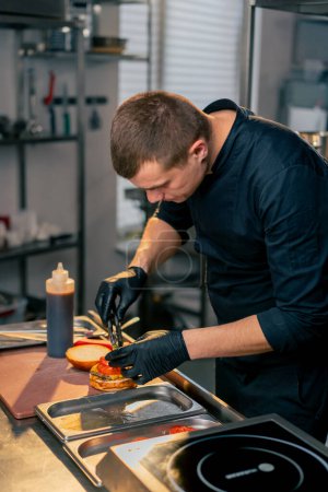 Foto de El chef en la cocina del establecimiento pone tomates en una hamburguesa casi terminada - Imagen libre de derechos