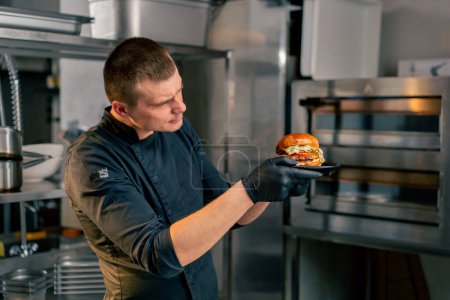 Photo for The chef in the kitchen of the establishment examines in his hands a ready-made burger lying on a black plate - Royalty Free Image