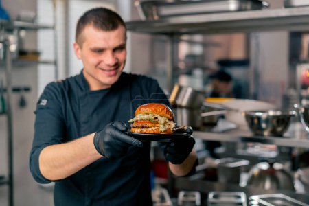 the chef in the kitchen of the establishment examines in his hands a ready-made burger lying on a black plate