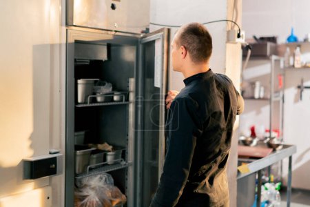 Foto de Un chef se para en una cocina profesional con un uniforme negro y mira a través de lo que está en la lista en el refrigerador - Imagen libre de derechos