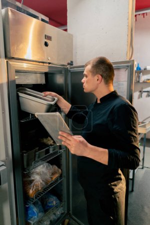Téléchargez les photos : Un chef se tient dans une cuisine professionnelle dans un uniforme noir et regarde à travers ce qui est sur la liste dans le réfrigérateur - en image libre de droit