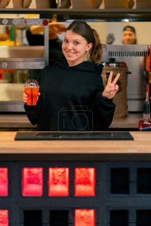 Foto de Una joven en un restaurante se para en el mostrador del bar con un cóctel en la mano y sonríe - Imagen libre de derechos