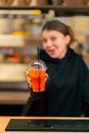 Photo for A young girl in a restaurant behind the bar holds out a cocktail to the camera and smiles - Royalty Free Image