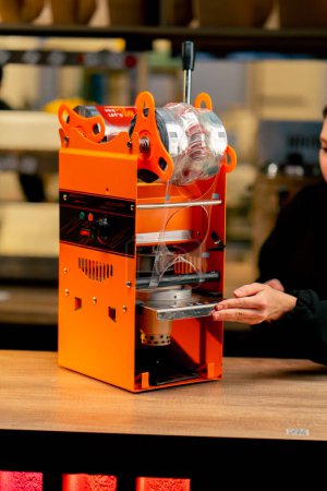 Nahaufnahme einer orangefarbenen Maschine zum Verpacken von Getränken mit Kunststoff auf einer Theke Hände beim Einrichten der Maschine