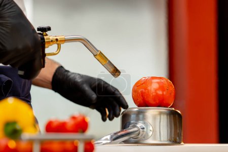 Foto de Primer plano, el chef se para en la cocina con guantes negros sostiene un quemador y quema un tomate - Imagen libre de derechos