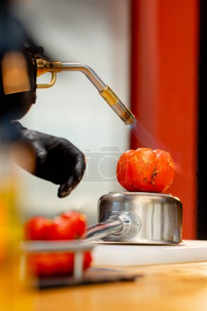 Foto de Primer plano, el chef se para en la cocina con guantes negros sostiene un quemador y quema un tomate - Imagen libre de derechos