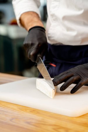 Foto de Primer plano de un chef manos en guantes negros rebanando queso en una mesa en la cocina - Imagen libre de derechos