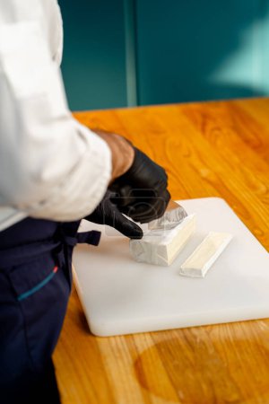 Téléchargez les photos : Gros plan d'un chef mains en gants noirs tranchant du fromage sur une table dans la cuisine - en image libre de droit