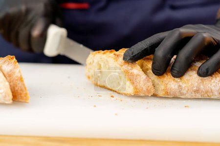 Foto de Primer plano de un chef manos en guantes negros rebanando baguettes en una mesa en una pizarra blanca - Imagen libre de derechos