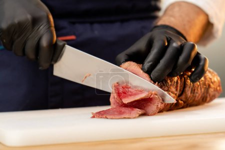 Foto de Primer plano de un chefs manos en guantes negros cortar un pedazo de carne seca cruda en un tablero - Imagen libre de derechos
