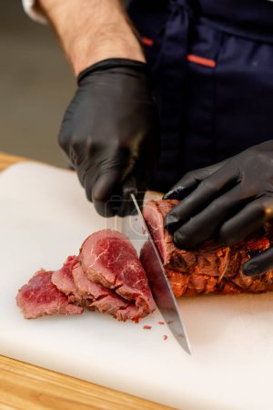 Foto de Primer plano de un chefs manos en guantes negros cortar un pedazo de carne seca cruda en un tablero - Imagen libre de derechos