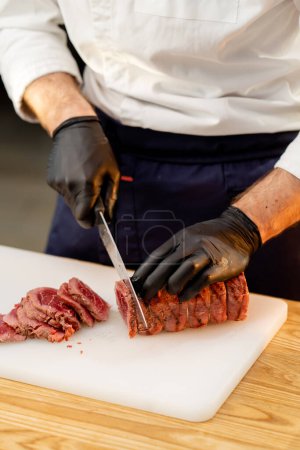 Photo for Close-up of a chefs hands in black gloves cutting a piece of raw jerky meat on a board - Royalty Free Image