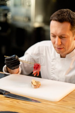 Photo for Close-up of the face of a chef in black gloves applying a piece of raw cured meat piece of baguette with tweezers - Royalty Free Image