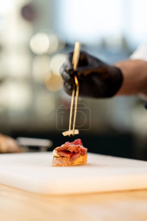 Foto de Primer plano de una mano de chefs en guantes negros colocando un pedazo de apio en un pedazo de baguette con pinzas - Imagen libre de derechos