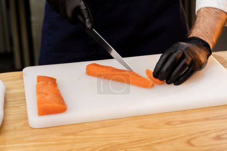 Foto de Primer plano de las manos de un chef con guantes negros cortando filete de salmón en una pizarra blanca - Imagen libre de derechos