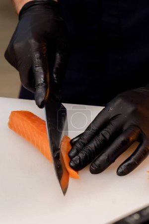 Foto de Primer plano de las manos de un chef con guantes negros cortando filete de salmón en una pizarra blanca - Imagen libre de derechos