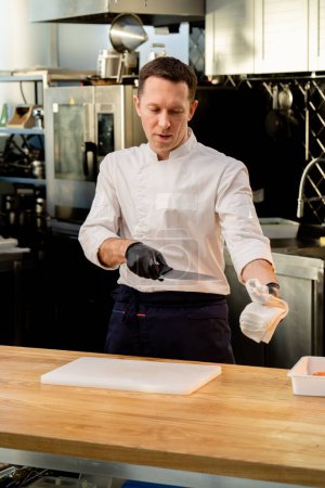 Photo for A chef in a white jacket in the kitchen wearing black gloves wipes knife with a white towel - Royalty Free Image