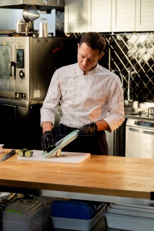 Foto de Un chef con una chaqueta blanca en la cocina con guantes negros corta pepinos antes de cocinar - Imagen libre de derechos