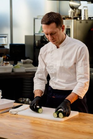 Foto de Un chef en una chaqueta blanca en la cocina con guantes negros pone pepinos antes de cocinar - Imagen libre de derechos